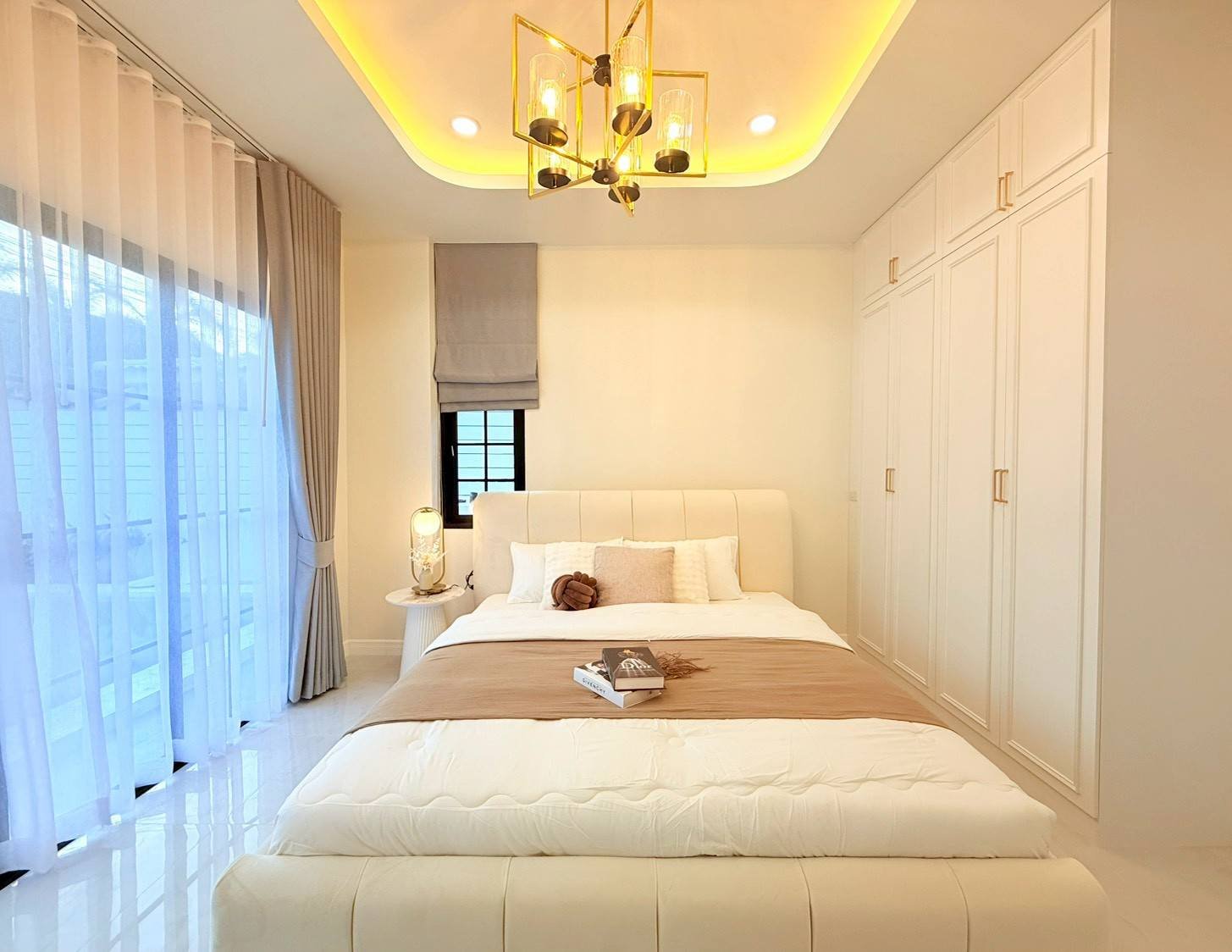 Pattaya_Tropical_2_Village_Dlh535_5 Bright modern bedroom with a white upholstered bed, beige headboard, and built-in white wardrobes on the right; sheer curtains over large windows.