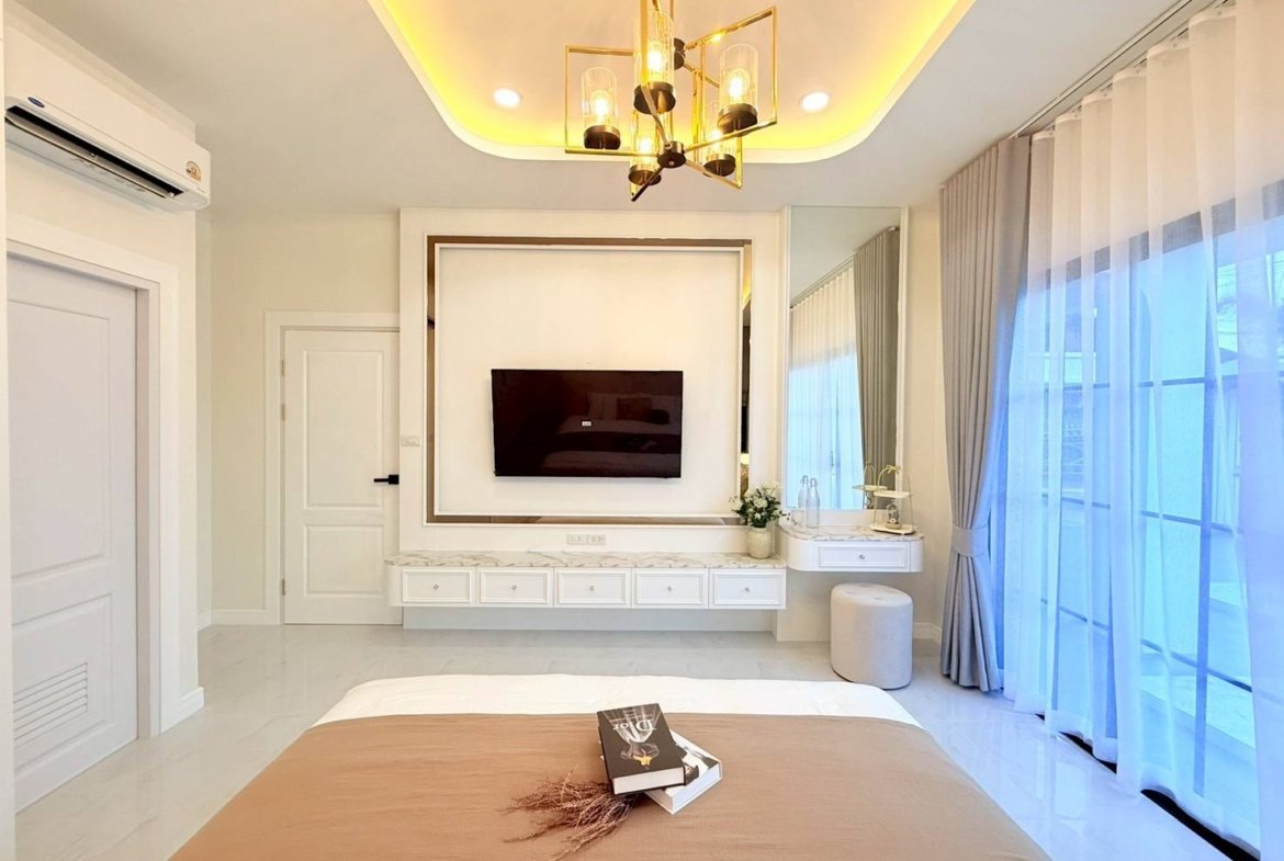 Bright hotel room with wall-mounted TV, marble vanity, and a large mirror framed by warm lighting above a bed side by side with sheer, floor-to-ceiling curtains.