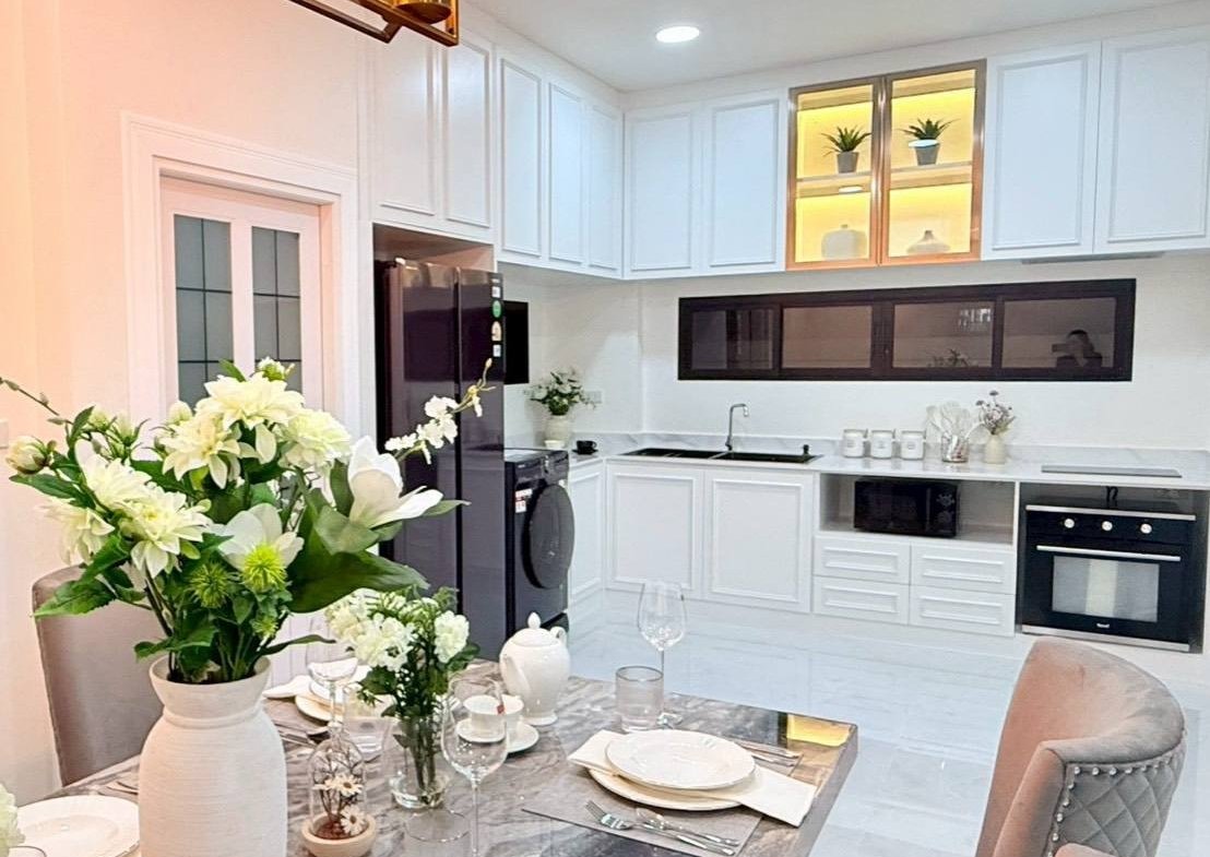 Modern white kitchen with built-in appliances and a dining table set for dinner, featuring a large white vase with flowers.