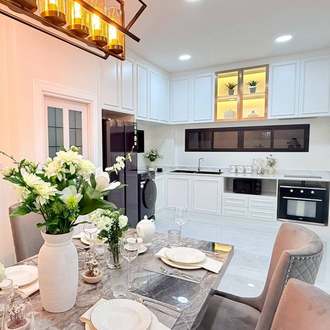 Pattaya_Tropical_2_Village_Dlh535_8 Modern white kitchen with built-in appliances and a dining table set for dinner, featuring a large white vase with flowers.