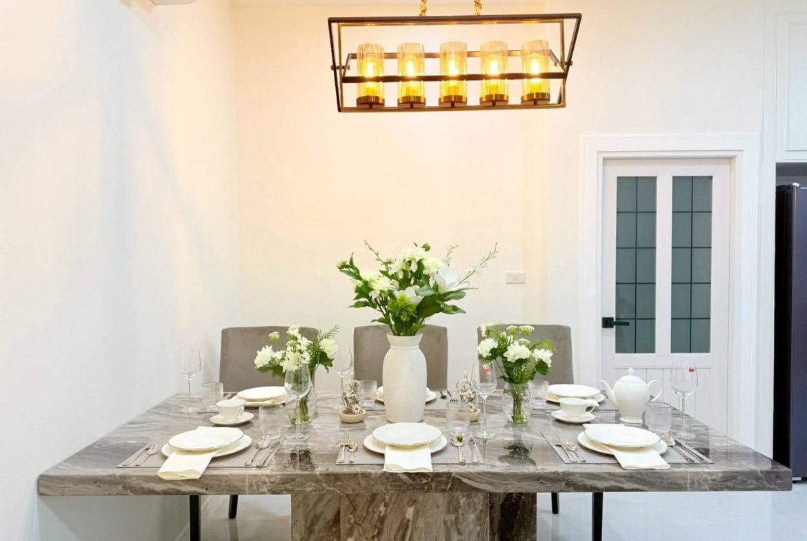 Marble dining table set for six with white plates, napkins, and glassware; central white vase with greenery and flowers under a warm chandelier.