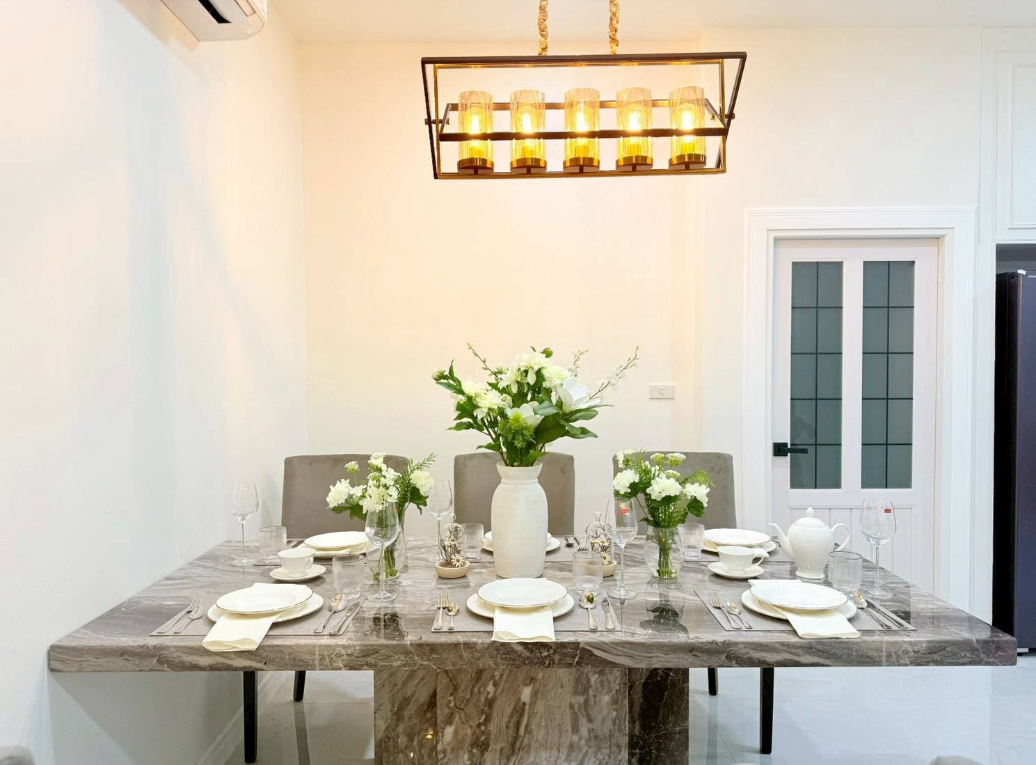 Pattaya_Tropical_2_Village_Dlh535_9 Marble dining table set for six with white plates, napkins, and glassware; central white vase with greenery and flowers under a warm chandelier.