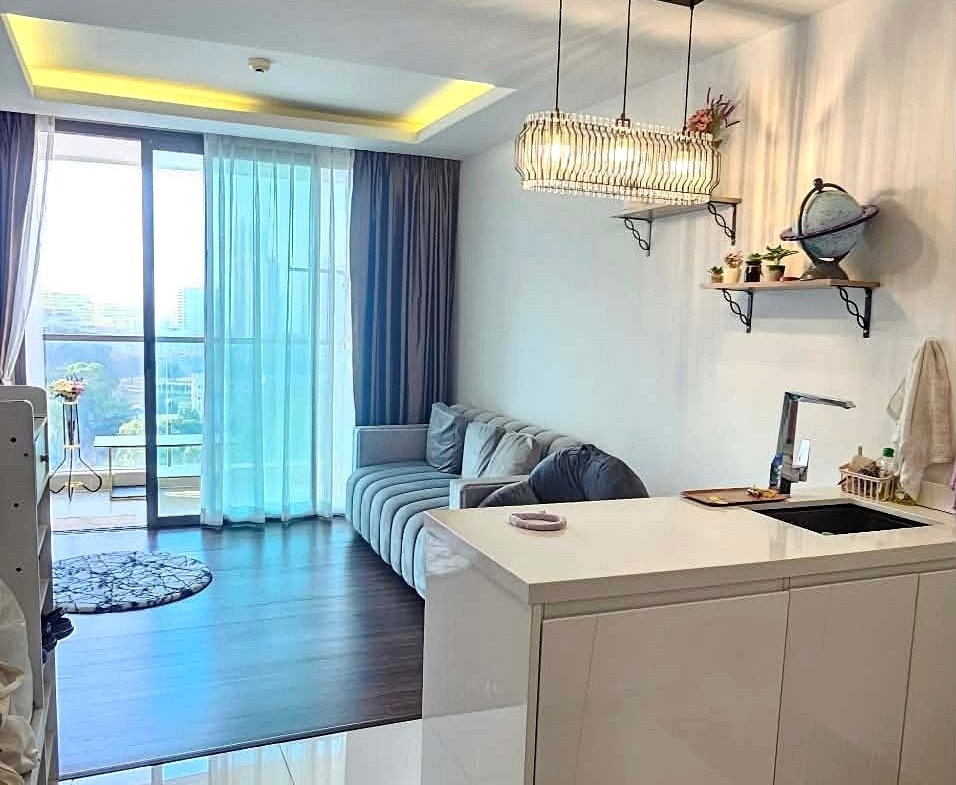 Open-plan living area with a white glossy kitchen island, gray sofa, and blue sheer curtains at sliding balcony doors.