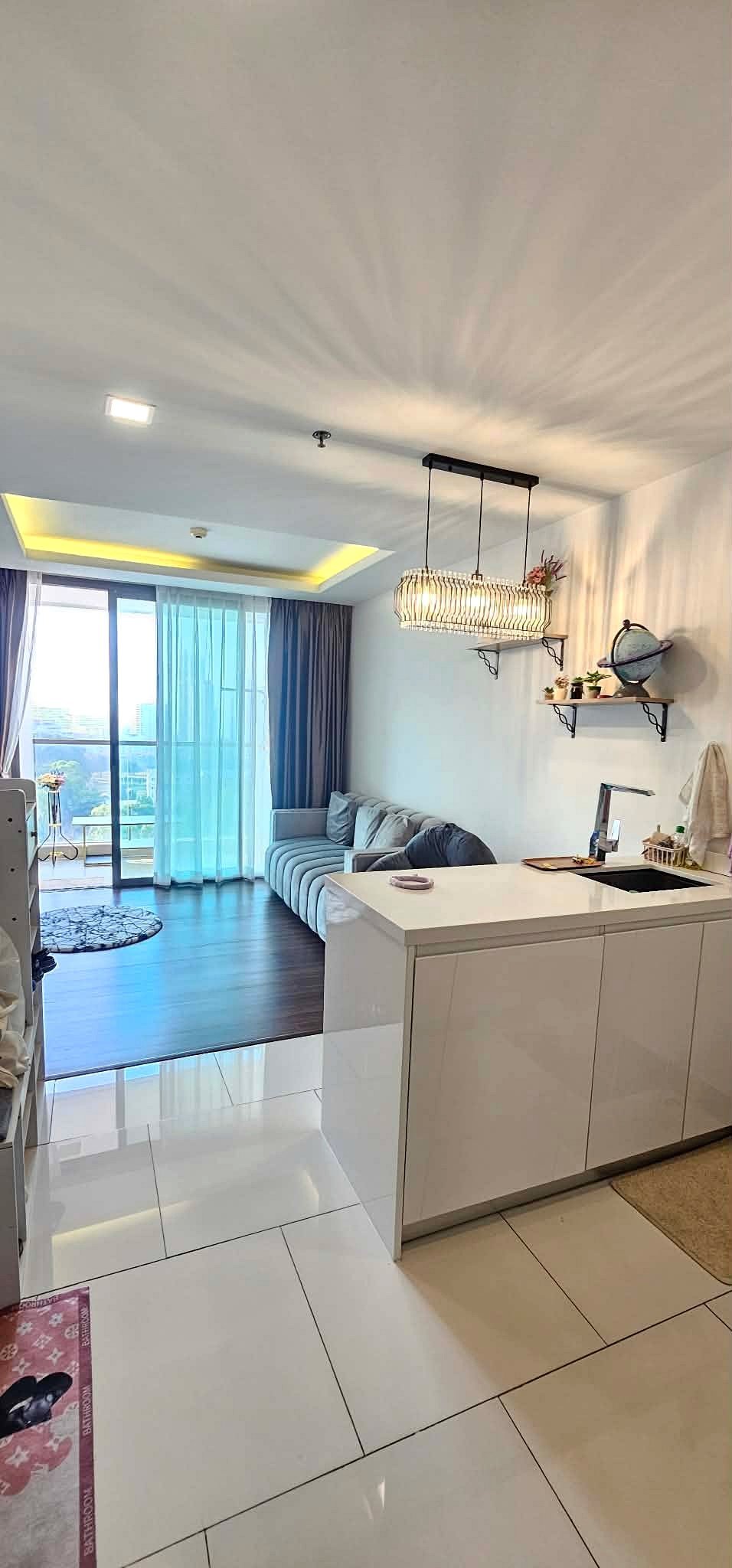 Peak_Tower_Sale_Rent_Dlc365_Rdl337_10 Open-plan living area with a white glossy kitchen island, gray sofa, and blue sheer curtains at sliding balcony doors.