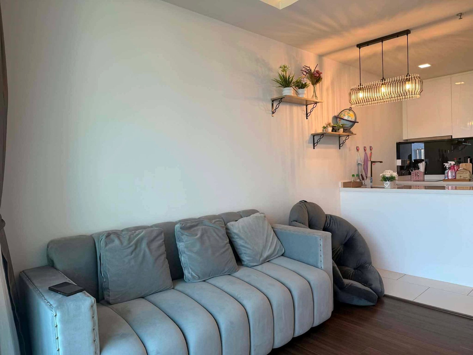 Peak_Tower_Sale_Rent_Dlc365_Rdl337_12 Light gray sectional sofa in a bright living area with wall shelves holding plants and a globe, and a metal cage pendant light overhead in the kitchen.