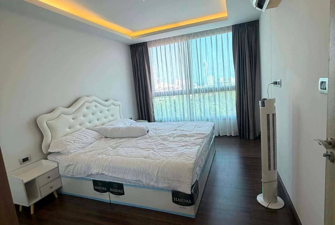 Hotel bedroom with a white tufted headboard bed, nightstand, and a large window with sheer curtains letting in light, plus ceiling ambient lighting.