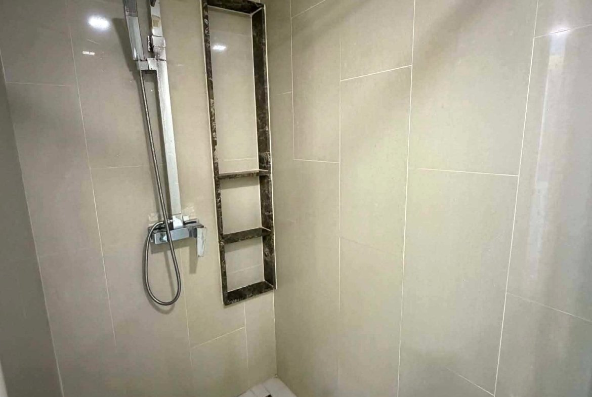 Modern shower area with square rainfall showerhead, handheld wand on a vertical rail, marble-framed square niche, beige tiles, and a white floor with a black mosaic border.