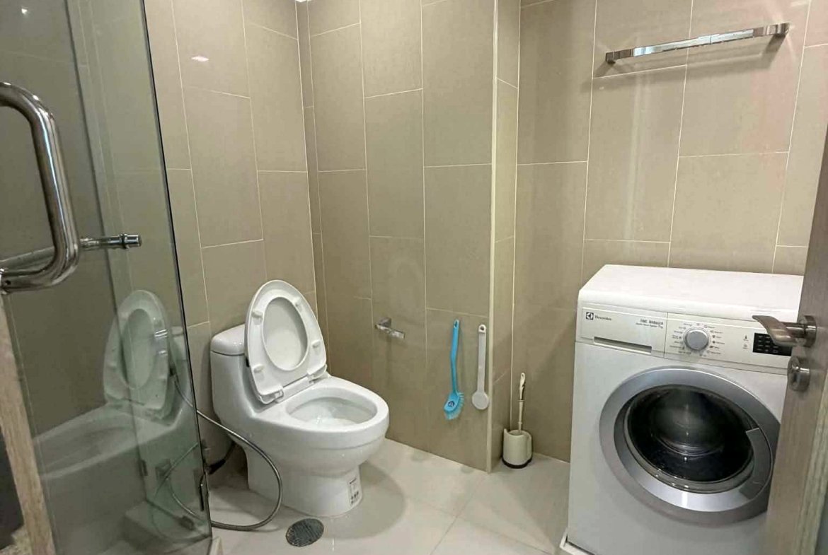 Compact beige-tiled bathroom with a glass shower enclosure on the left, a white toilet, and a front-loading washing machine on the right.