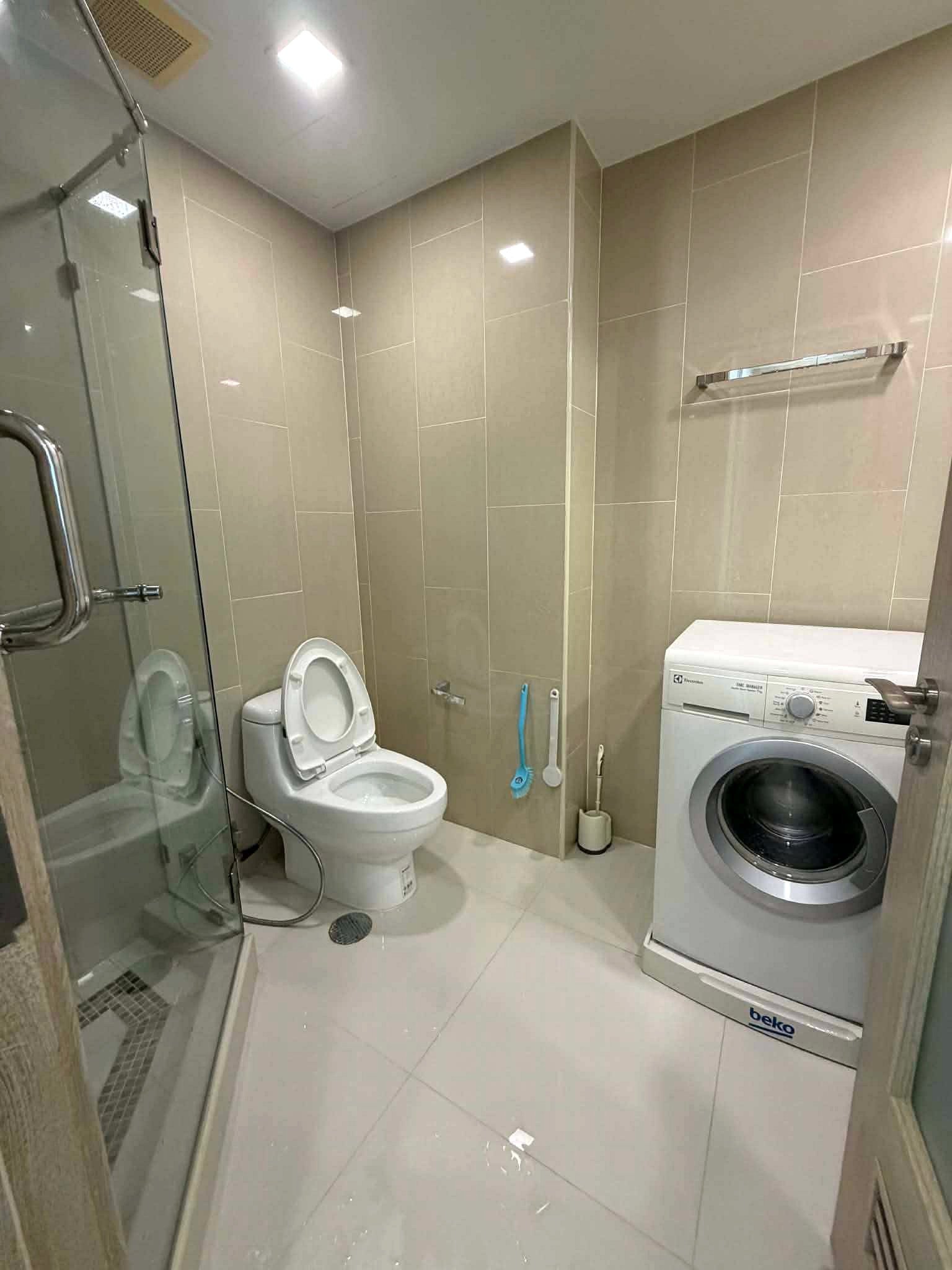 Peak_Tower_Sale_Rent_Dlc365_Rdl337_16 Compact beige-tiled bathroom with a glass shower enclosure on the left, a white toilet, and a front-loading washing machine on the right.