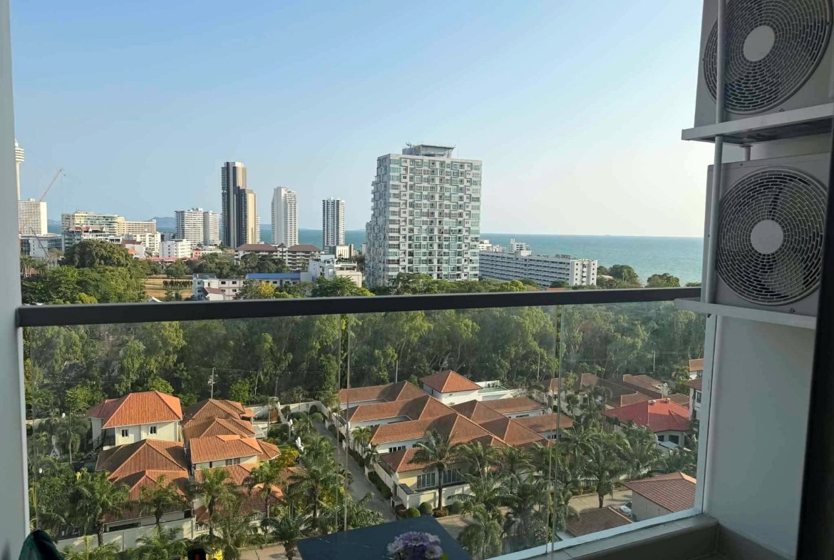 Coastal city view from a balcony: mid‑rise buildings, red-tiled houses, palm trees, and the sea on the horizon.