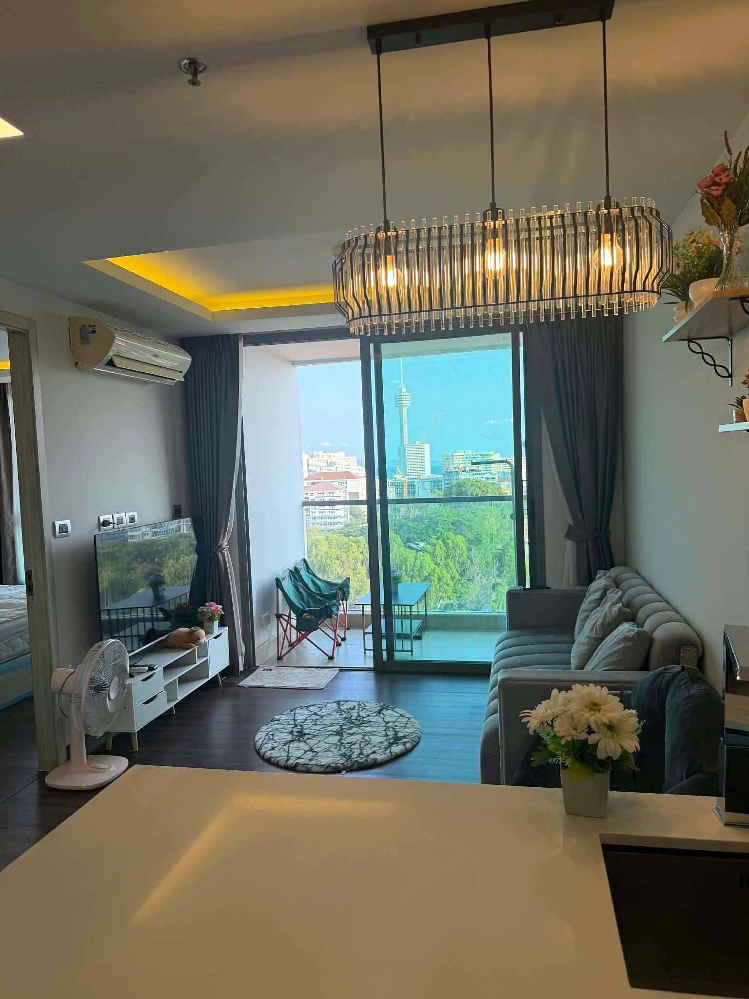 Peak_Tower_Sale_Rent_Dlc365_Rdl337_3 Modern living room with a metal chandelier over sliding balcony doors and a city skyline view, gray sofa and flowers on a white counter in the foreground.