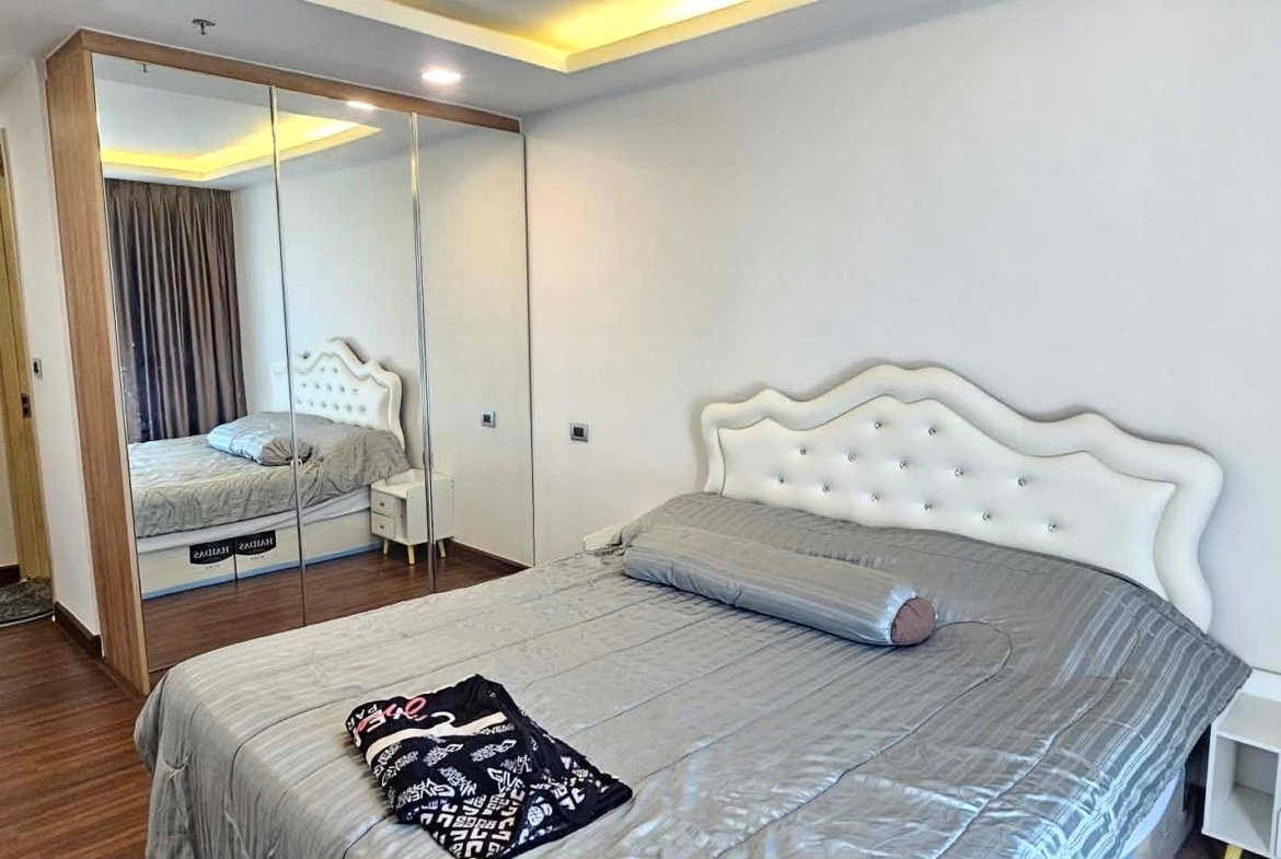 Bedroom with a tufted white headboard, gray bedding, and white nightstand on a wooden floor; large mirrored wardrobe reflects the bed.