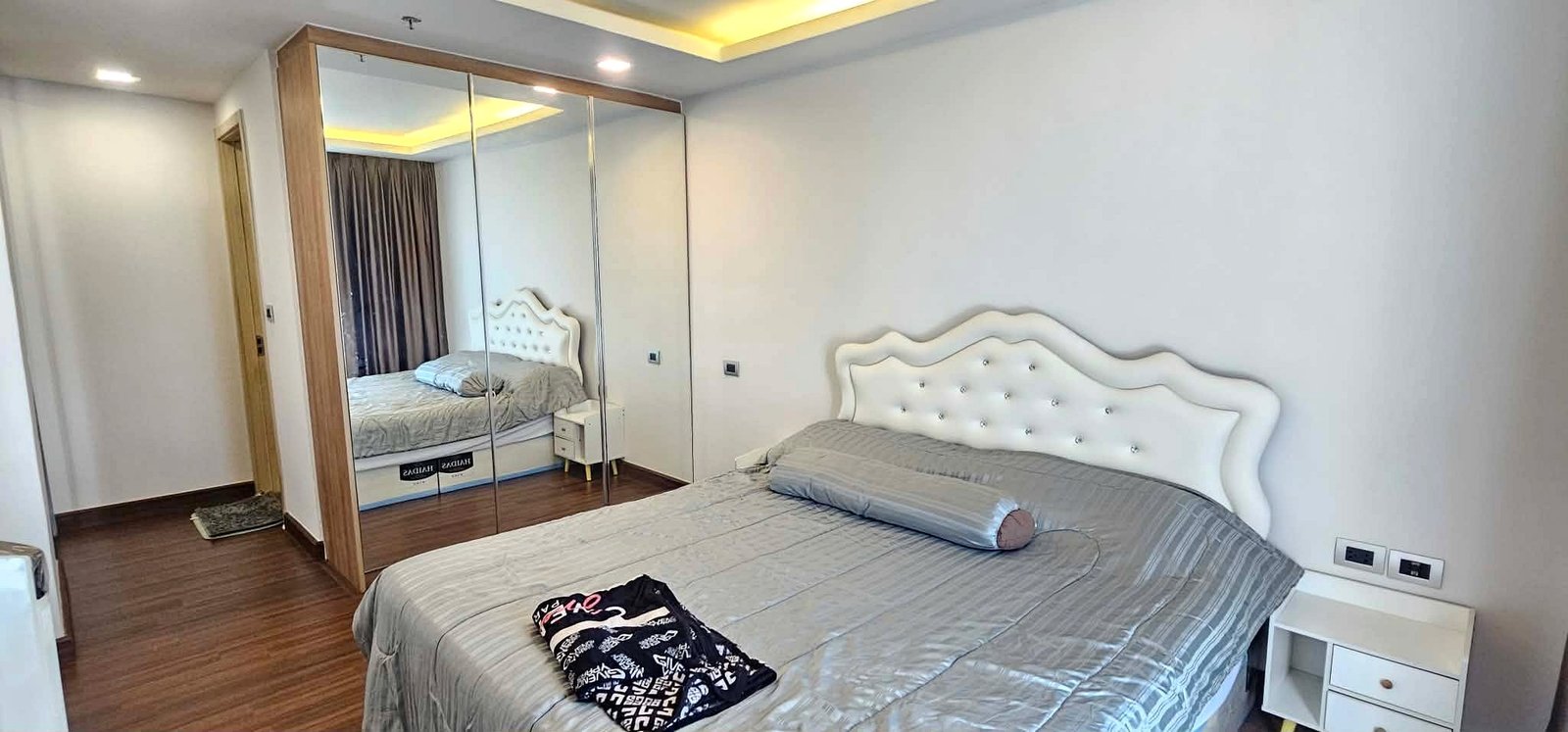 Peak_Tower_Sale_Rent_Dlc365_Rdl337_5 Bedroom with a tufted white headboard, gray bedding, and white nightstand on a wooden floor; large mirrored wardrobe reflects the bed.