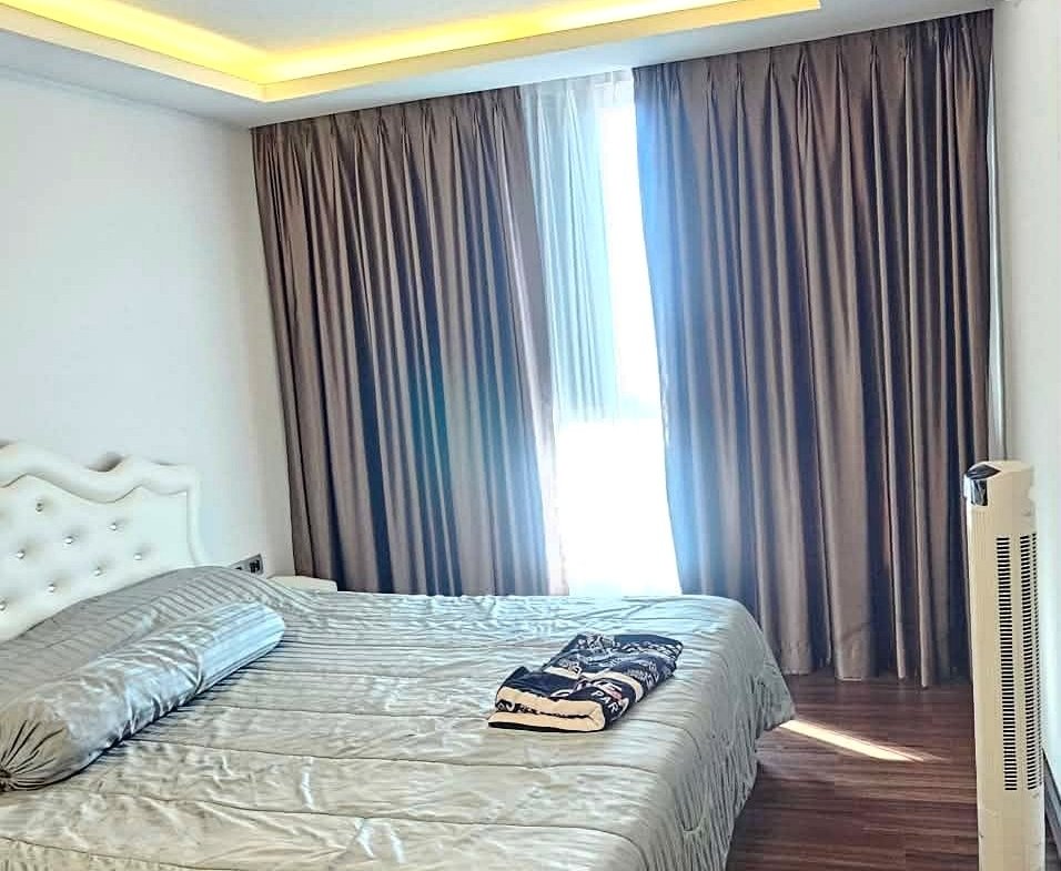 Hotel-style bedroom with a king bed, gray bedding, and a white tufted headboard; folded towels rest on the bed, dark curtains, and a wooden floor from ceiling cove lighting above.