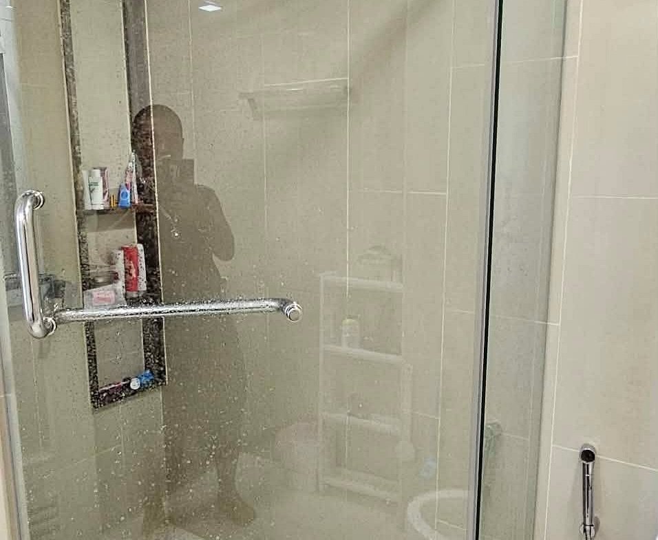 Glass shower enclosure with chrome hardware, tiled walls, and a built-in shelf visible inside; photographer’s silhouette reflected.