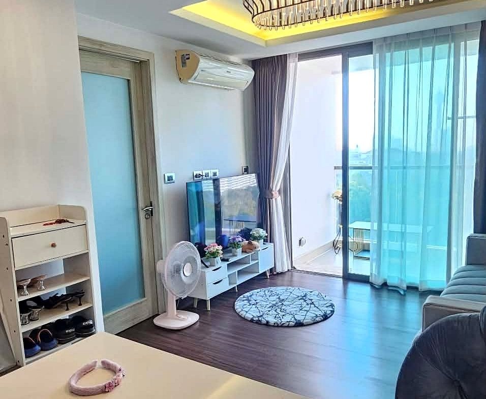 Modern living area with a white island in the foreground, a sink to the right, TV stand, fan, and balcony doors with blue sheer curtains.