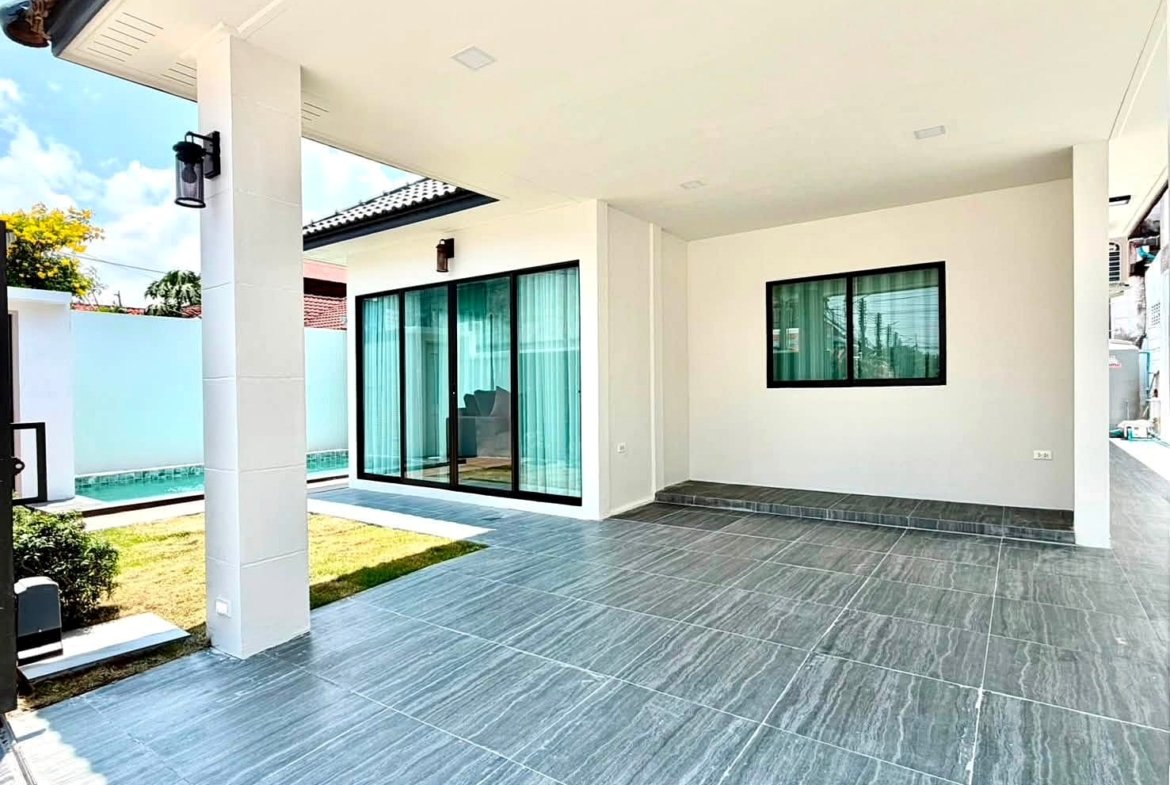 Modern house patio with gray tile flooring, white walls, and large glass sliding doors opening to the interior. A small lawn and privacy wall are visible in the background, with a pool edge along the side.