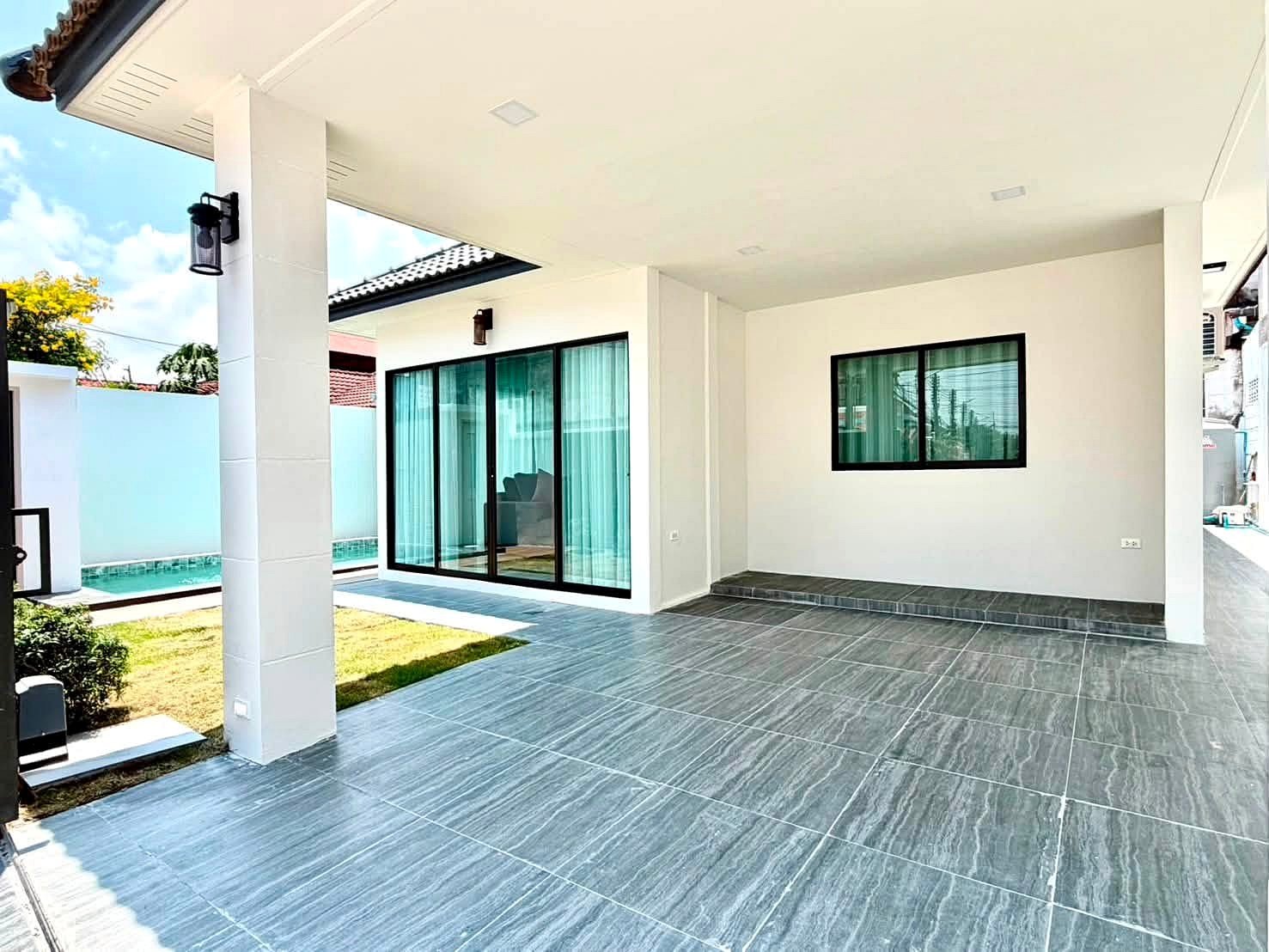 Modern house patio with gray tile flooring, white walls, and large glass sliding doors opening to the interior. A small lawn and privacy wall are visible in the background, with a pool edge along the side.