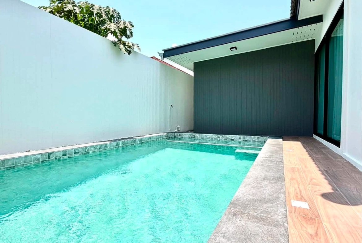 Modern rectangular backyard pool with turquoise water, white privacy wall, gray coping, and a wooden deck beside a dark gray wall and glass doors.