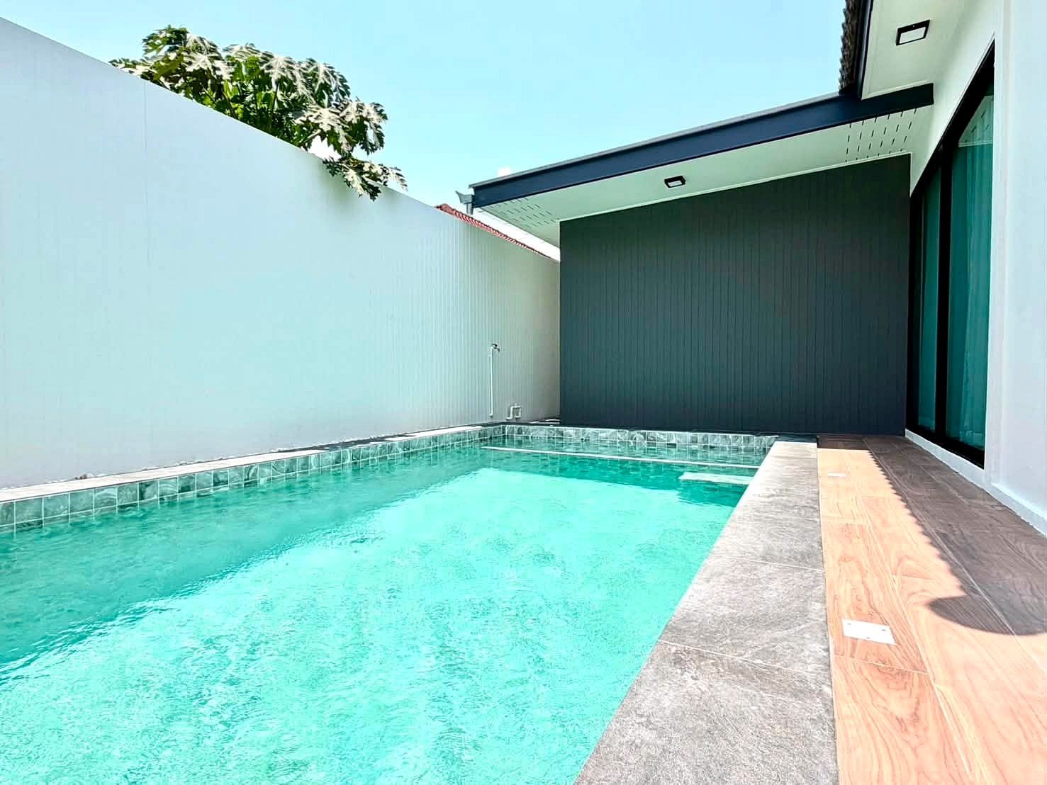 Modern rectangular backyard pool with turquoise water, white privacy wall, gray coping, and a wooden deck beside a dark gray wall and glass doors.