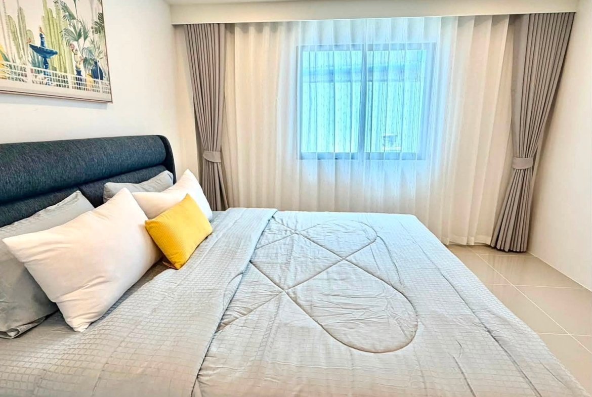 Bedroom with a large bed, gray quilted bedding, white pillows, a yellow accent pillow, and a window with sheer curtains.