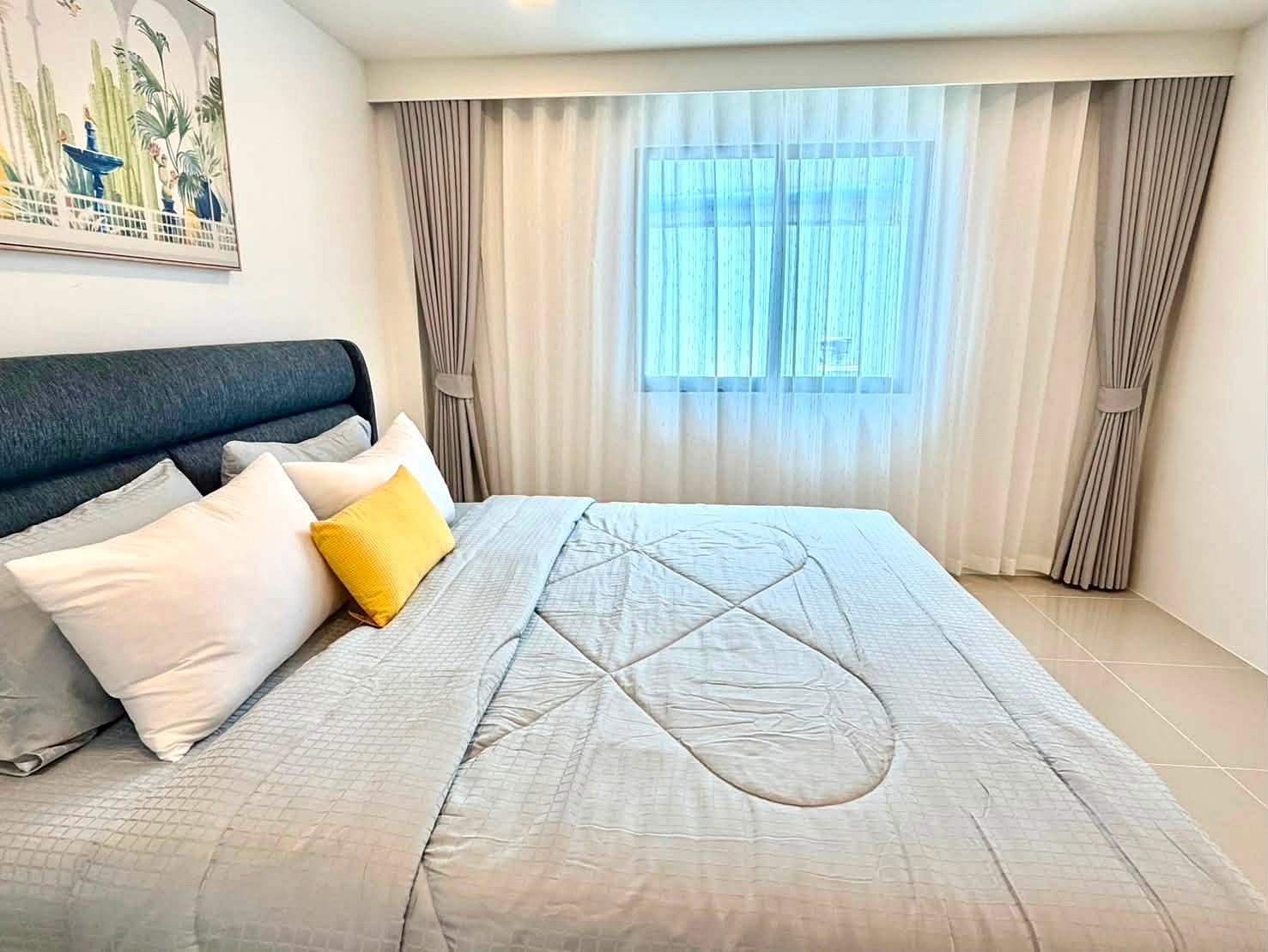 Bedroom with a large bed, gray quilted bedding, white pillows, a yellow accent pillow, and a window with sheer curtains.