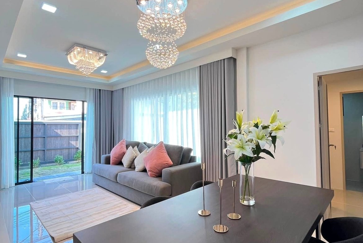 Modern living room with a crystal chandelier, grey sofa, and sheer-curtained windows by a glass door.