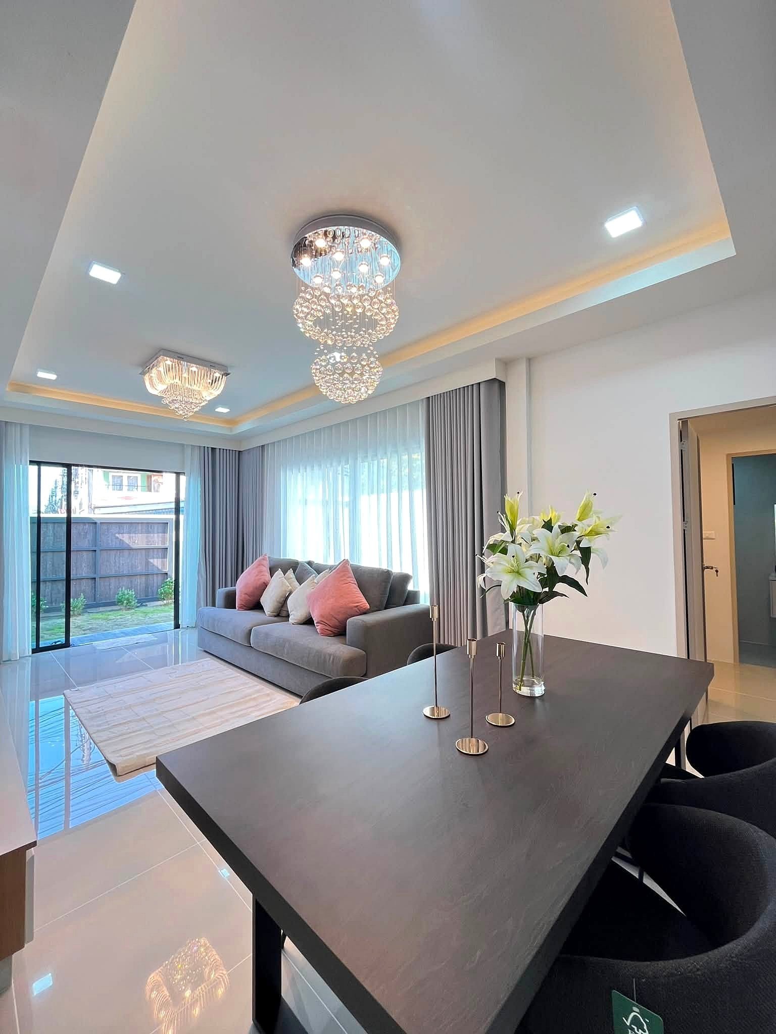 Modern living room with a crystal chandelier, grey sofa, and sheer-curtained windows by a glass door.