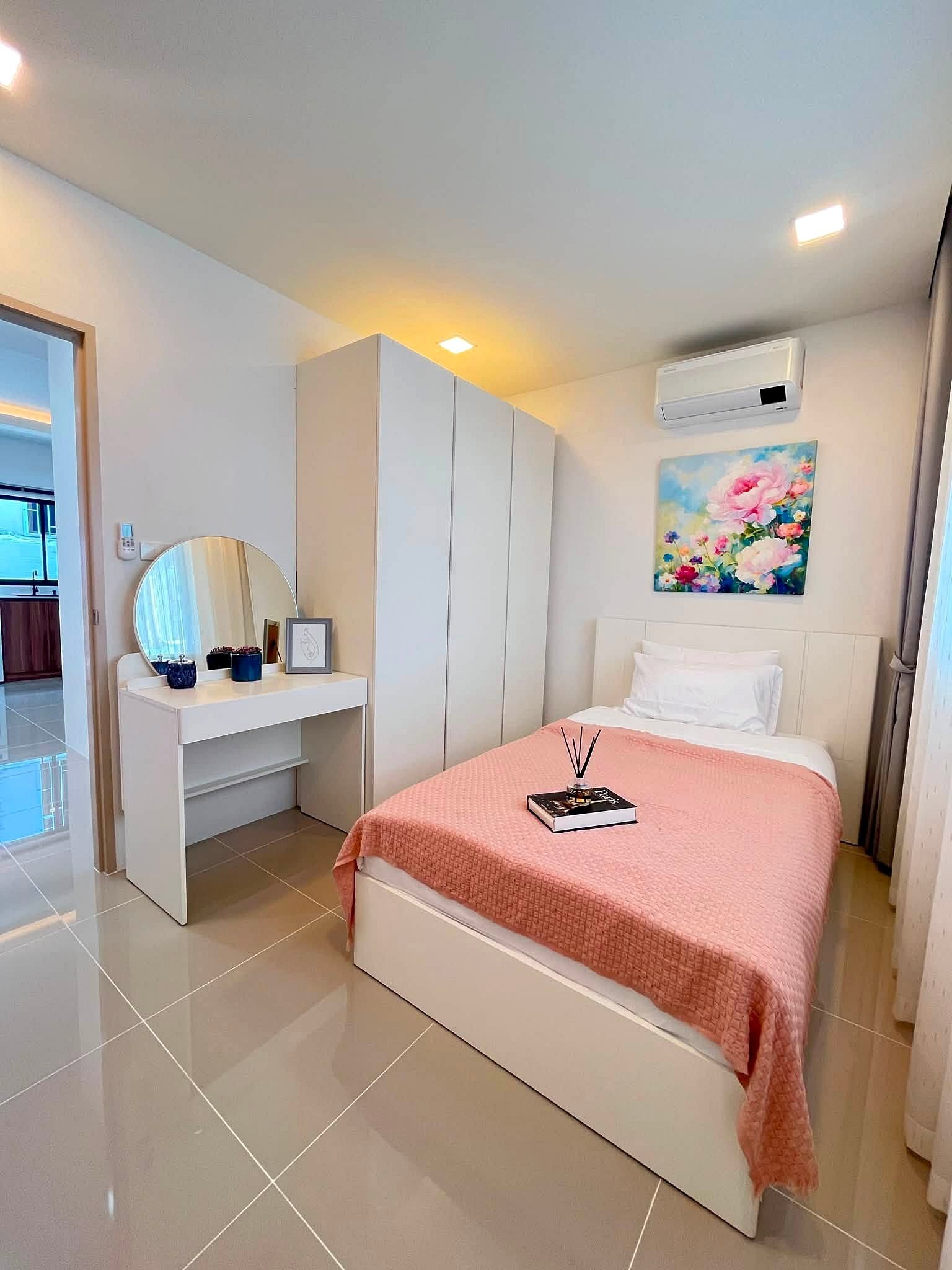 Bright contemporary bedroom with a pink quilt, white bed and built-in wardrobes; a dresser with a round mirror and blue decor accents along the wall art above the bed.