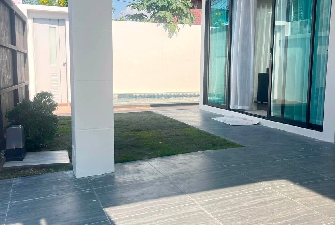 Modern home patio with large glass sliding doors, white walls, a white pillar, grey tiled floor, and hanging lanterns; a small pool and lawn beyond.
