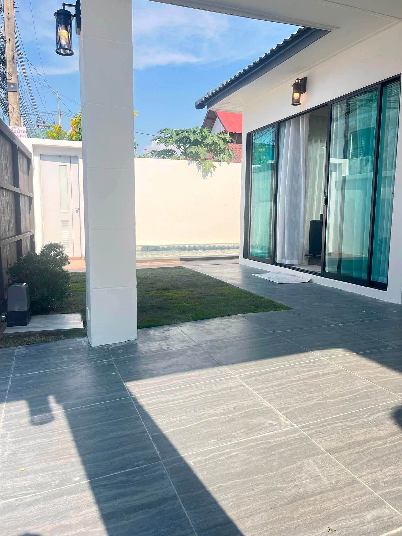 Modern home patio with large glass sliding doors, white walls, a white pillar, grey tiled floor, and hanging lanterns; a small pool and lawn beyond.