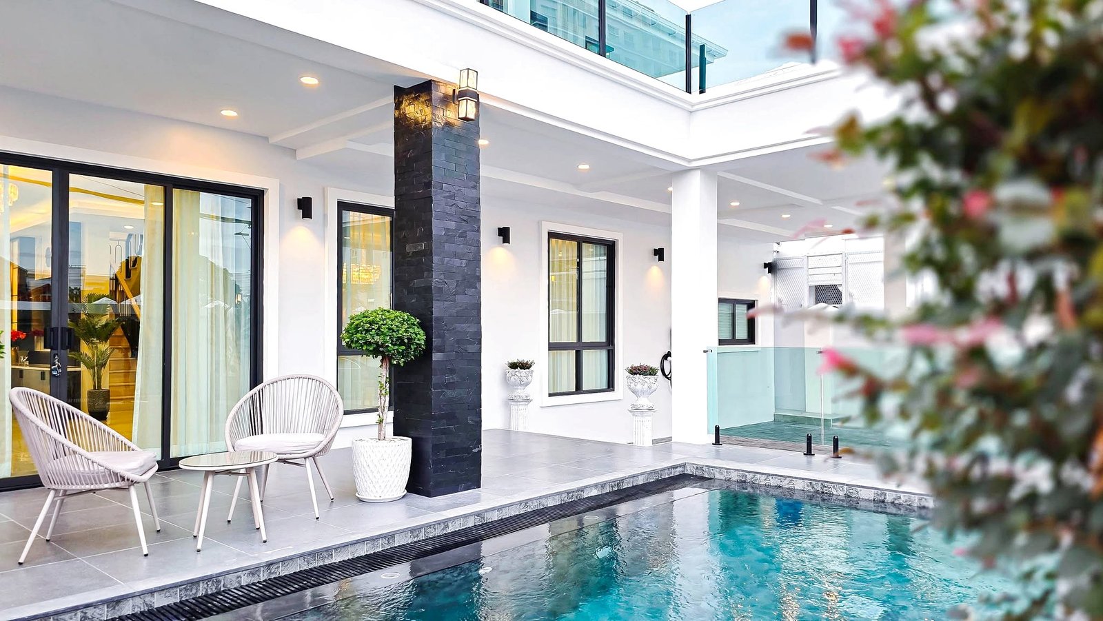 Modern poolside patio with white walls, black stone pillar, glass doors, and outdoor seating by the pool.