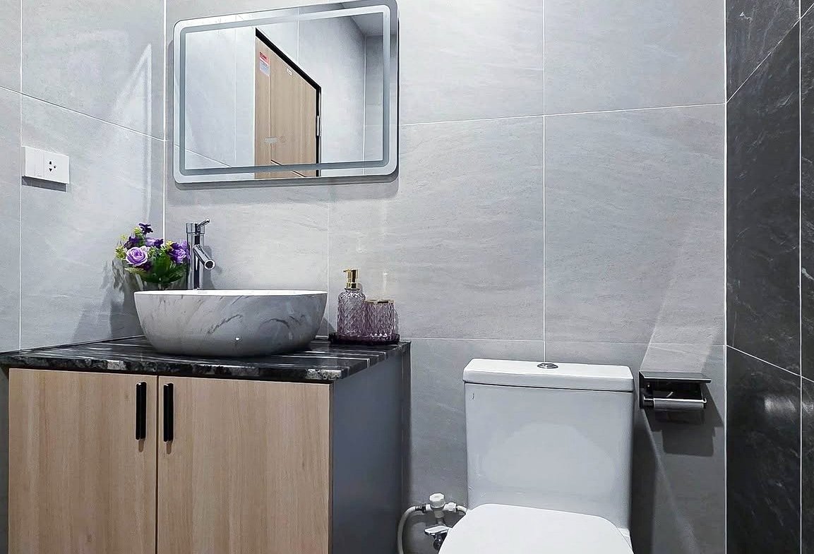 Modern bathroom with wooden vanity, marble vessel sink, mirror, and white toilet in a gray tiled room.