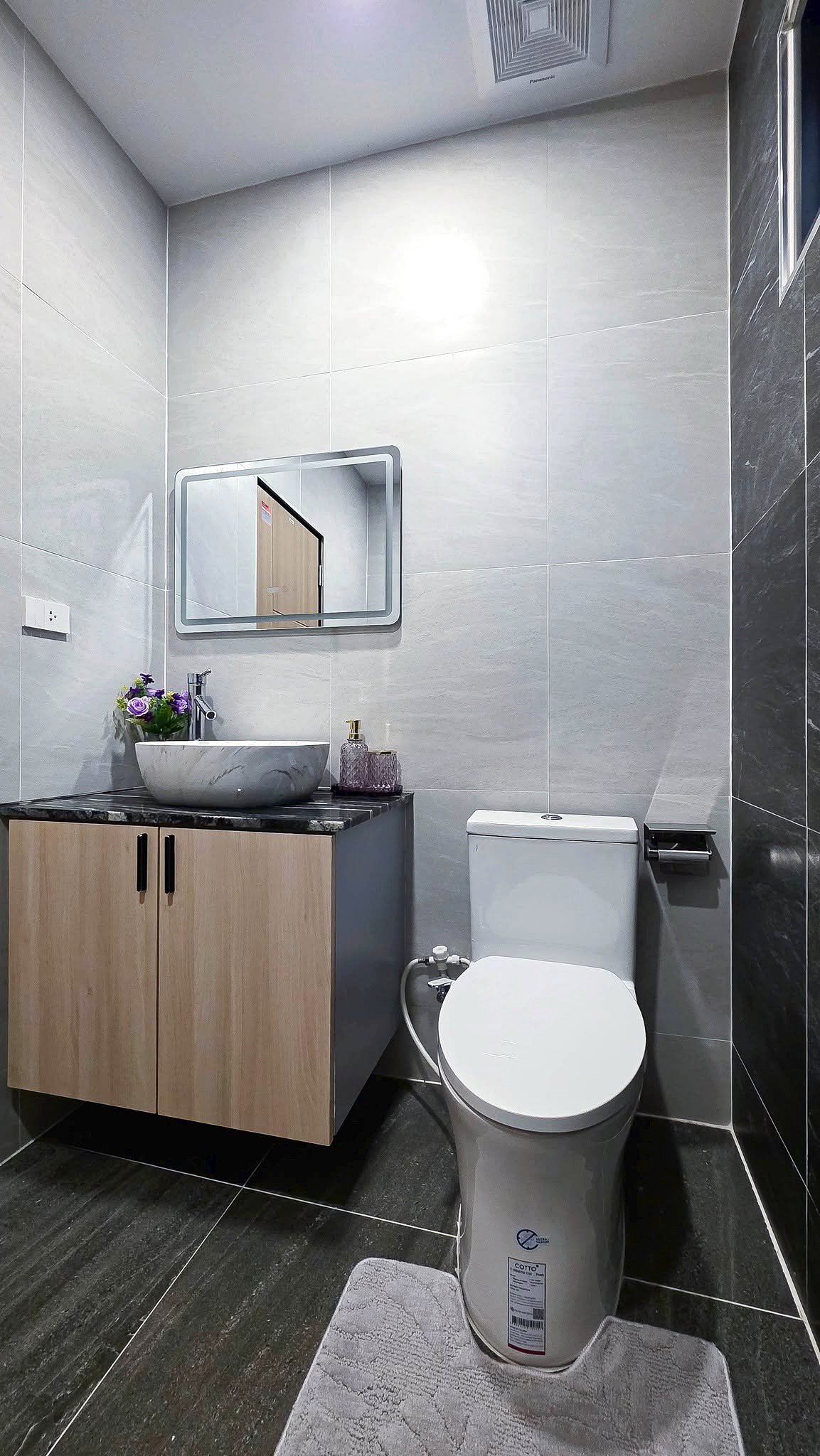 Modern bathroom with wooden vanity, marble vessel sink, mirror, and white toilet in a gray tiled room.