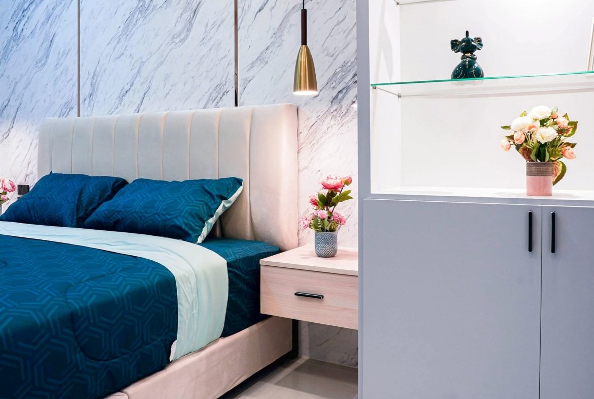 Modern bedroom with navy blue bedding, a white padded headboard, marble wall panels, and a dresser with pink flowers on the right.