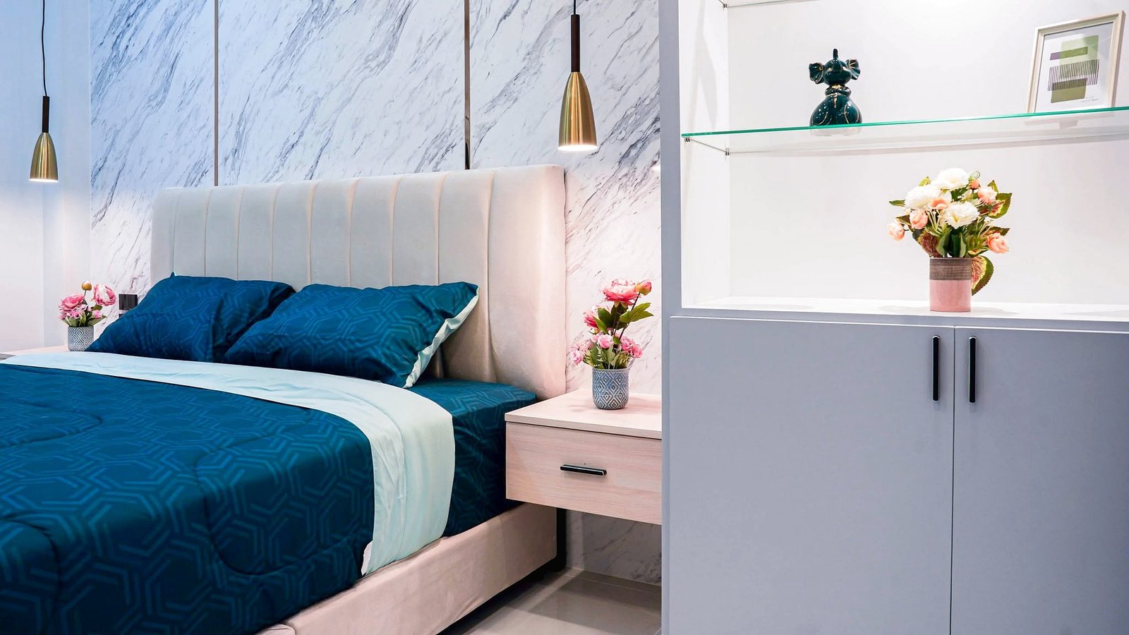 Modern bedroom with navy blue bedding, a white padded headboard, marble wall panels, and a dresser with pink flowers on the right.