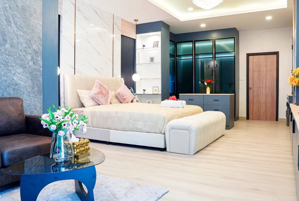 Modern hotel bedroom with a tufted beige bed, pink pillows, blue accent walls, and glass display shelves behind the headboard.