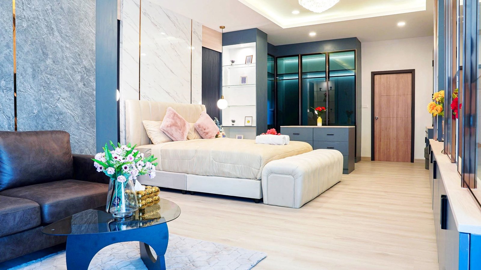Modern hotel bedroom with a tufted beige bed, pink pillows, blue accent walls, and glass display shelves behind the headboard.