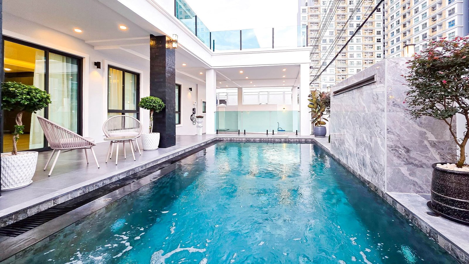 Modern poolside courtyard of a contemporary building with white walls, wicker chairs, and potted trees along a rectangular pool.