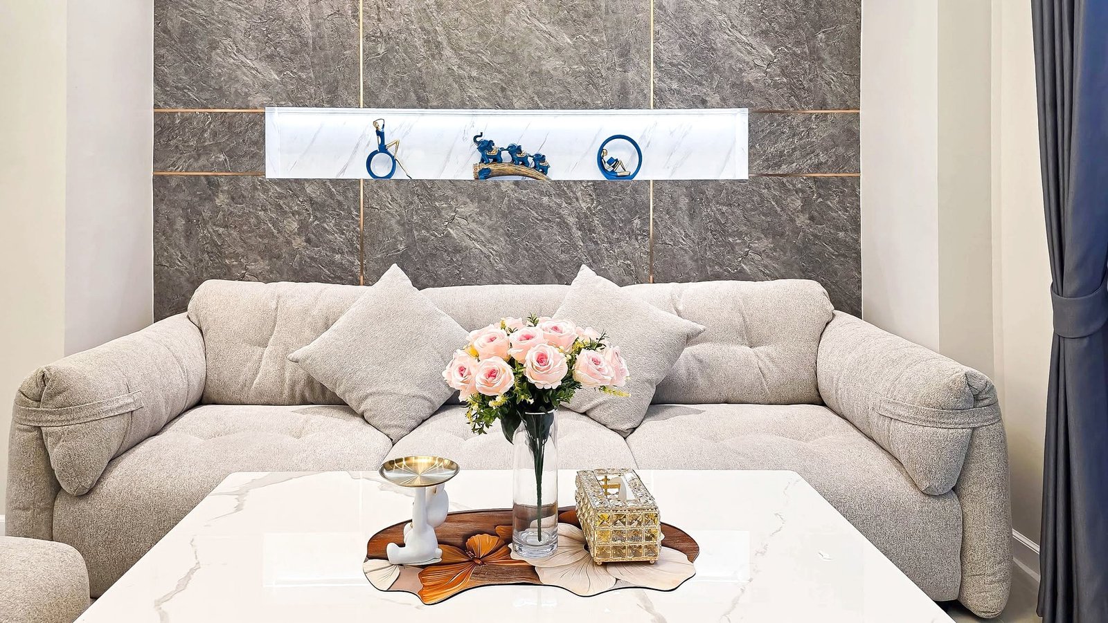 Light gray sectional sofa with cushions in a living room, a marble-top coffee table, and a vase of pink roses as center decor.