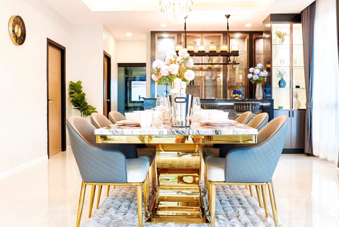 Luxurious dining room with a marble-topped table, blue quilted chairs, gold legs, and a chandelier above; bar area in the background.
