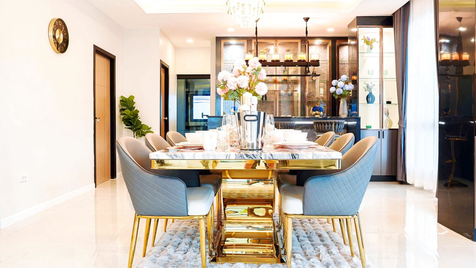 Luxurious dining room with a marble-topped table, blue quilted chairs, gold legs, and a chandelier above; bar area in the background.