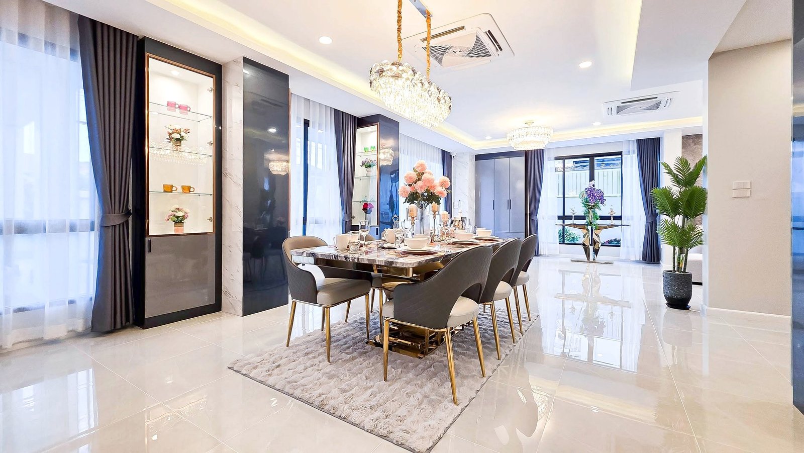 Luxurious dining room with a marble table, gold-legged chairs, crystal chandelier, and large windows with dark curtains.