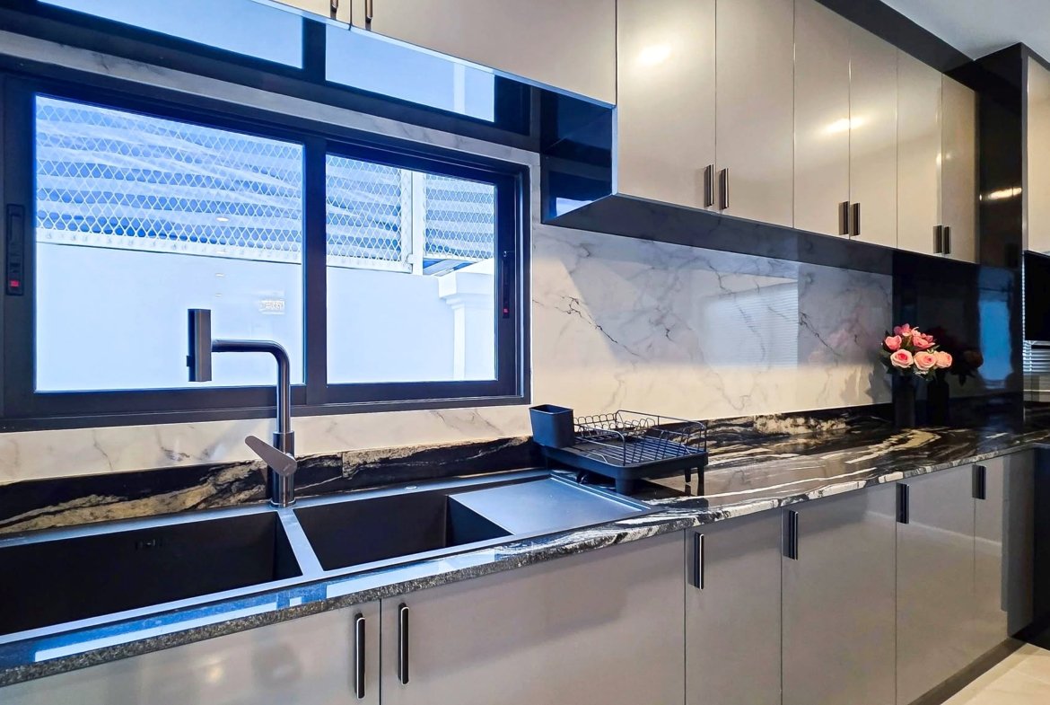 Modern kitchen with gray glossy cabinets, marble countertops, a double sink with chrome faucet, a dish rack, and a vase of pink roses on the counter near the window.