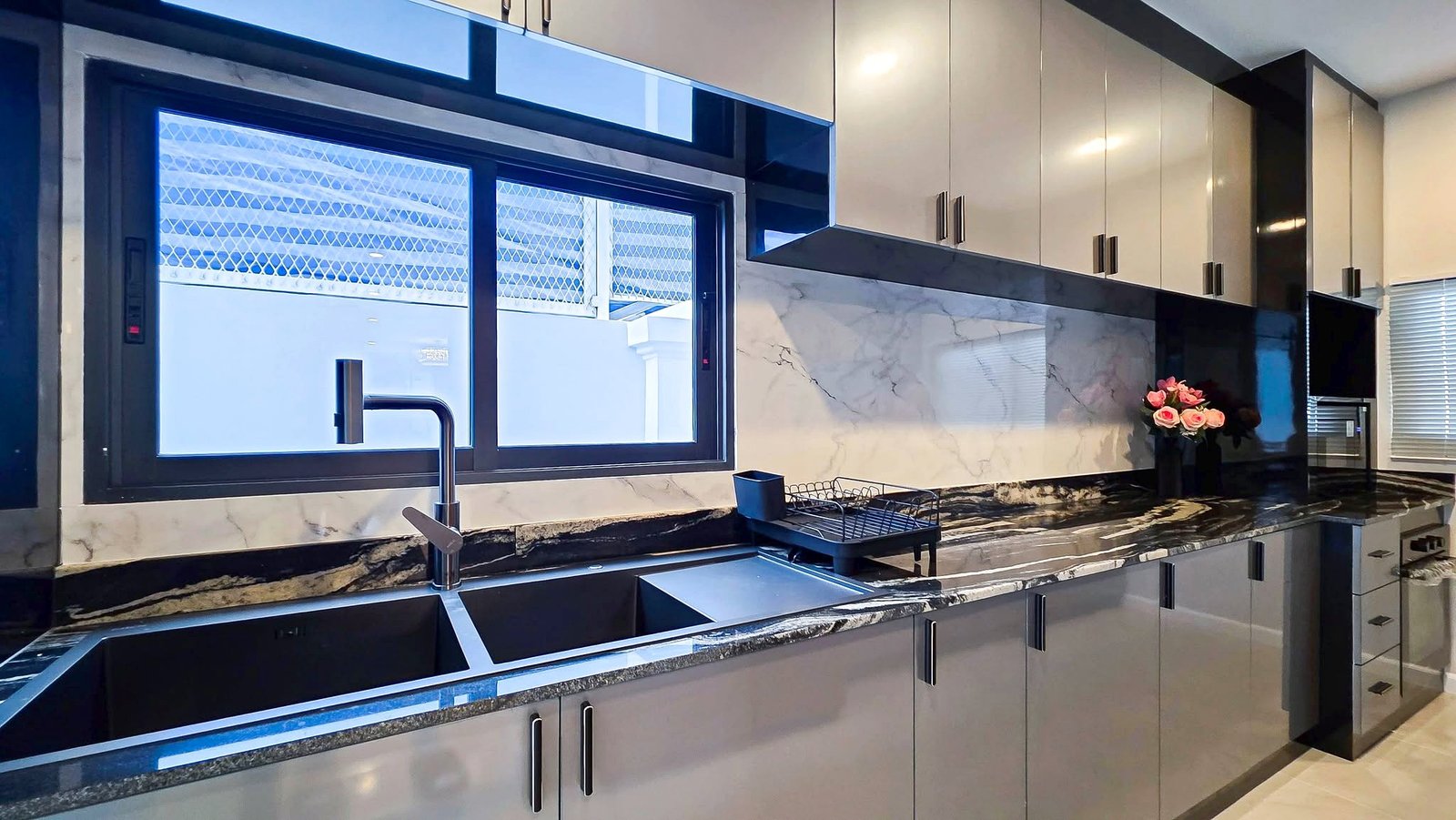 Modern kitchen with gray glossy cabinets, marble countertops, a double sink with chrome faucet, a dish rack, and a vase of pink roses on the counter near the window.