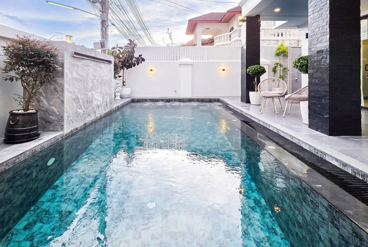 Rectangular backyard pool with turquoise water, gray stone deck, and white walls with potted plants and seating area nearby, in a modern courtyard.