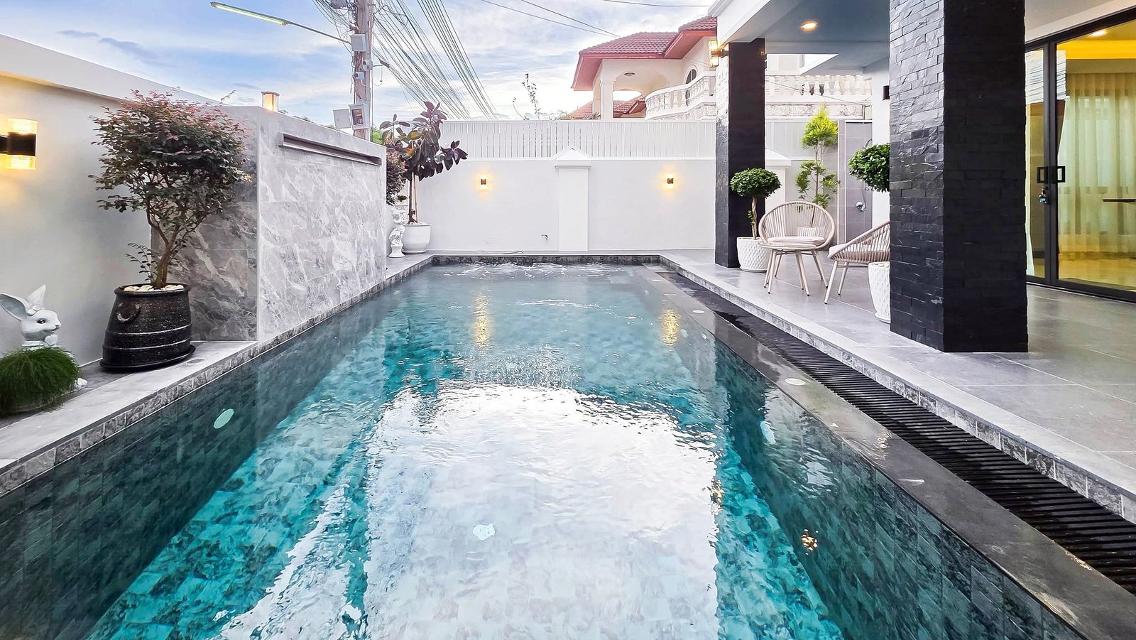 Rectangular backyard pool with turquoise water, gray stone deck, and white walls with potted plants and seating area nearby, in a modern courtyard.