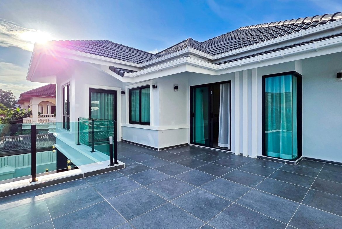Modern white house terrace with dark tile flooring, glass railing, and sliding doors under a sunny blue sky.