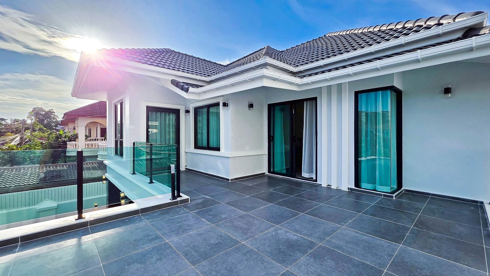 Modern white house terrace with dark tile flooring, glass railing, and sliding doors under a sunny blue sky.