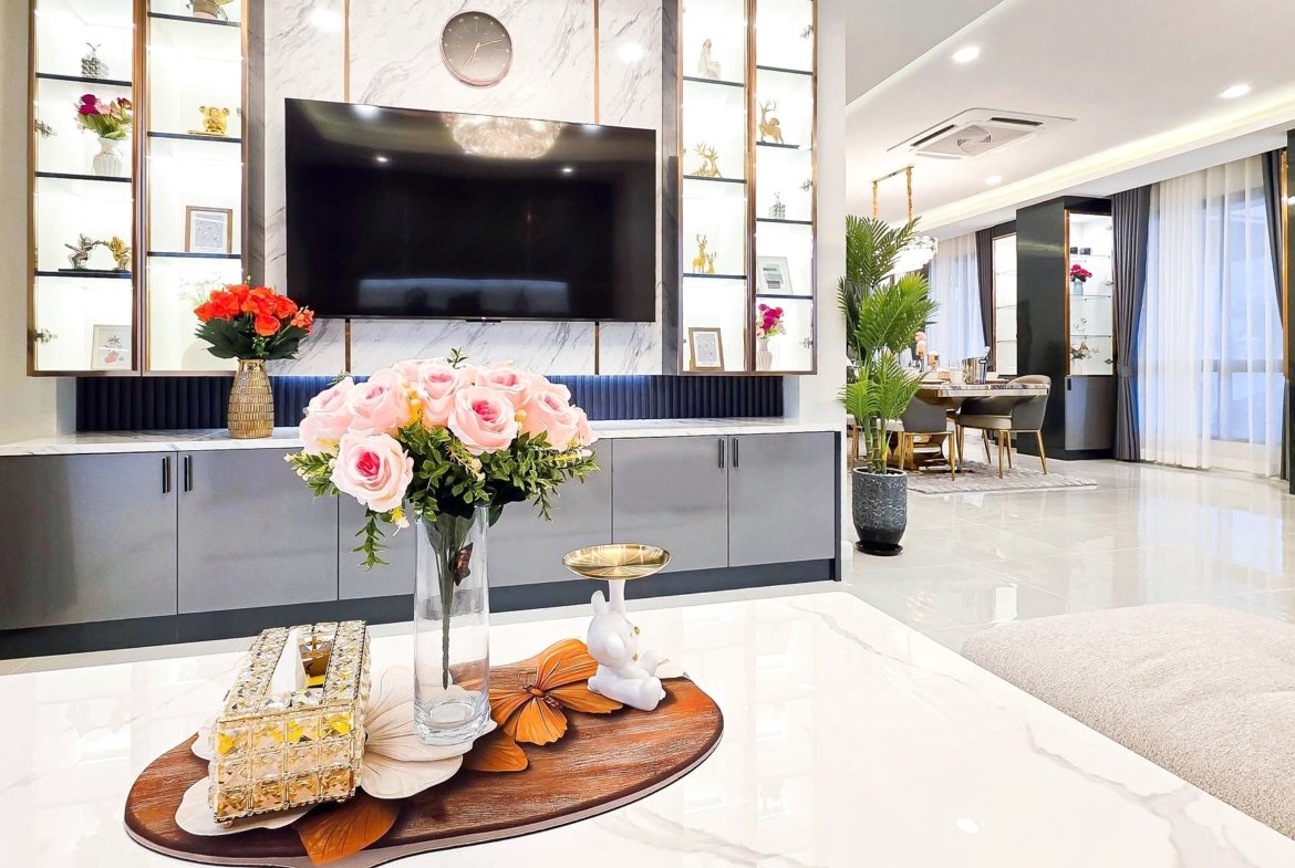 Bright modern living room with a large wall-mounted TV, marble accents, and glass display shelves filled with decor items.